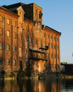 A red-bricked factory by a river. It’s empty and in a state of disrepair with broken windows.