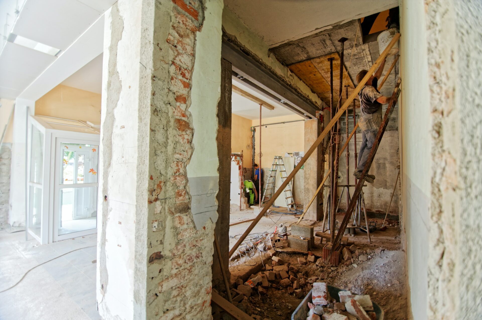 Interiors of a building undergoing dilapidations work. A pillar of bricks splits the image. To the left, a renovated entrance. To the right, two builders conduct repairs on the walls and ceiling.